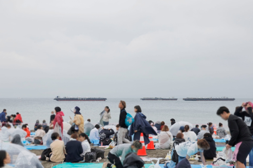 雨の中、多くの人々が会場であるビーチに集まる。海上に浮かんでいるのが打ち上げ場所の台船